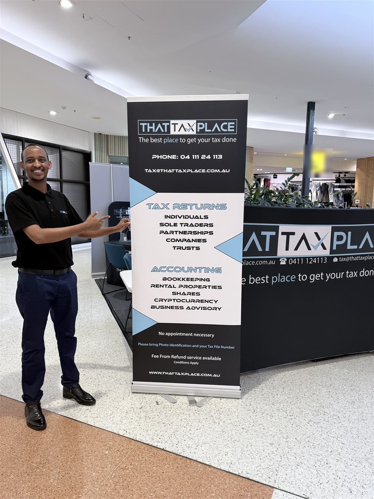 Naafac pointing to the That Tax Place banner at the Marketplace Beenleigh kiosk