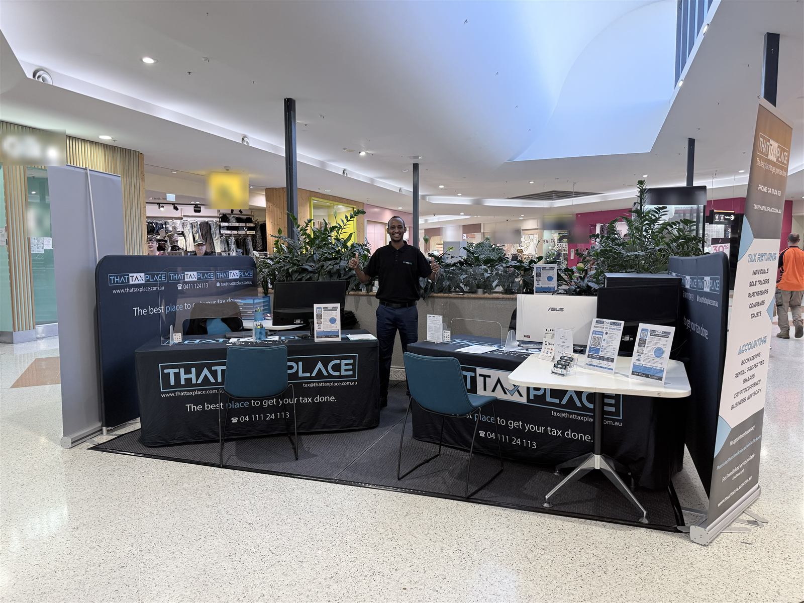 Naafac standing at the That Tax Place tax-season kiosk at Marketplace Beenleigh, with two client desks and branded signage